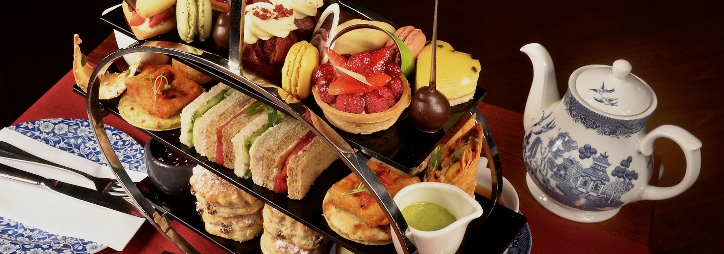 A tiered afternoon tea tray with sandwiches, scones, pastries, and a blue-and-white teapot on a table.