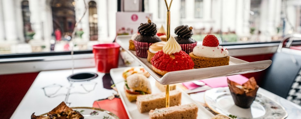 Tiered afternoon tea trays with sandwiches and cakes beside a bus window in London.