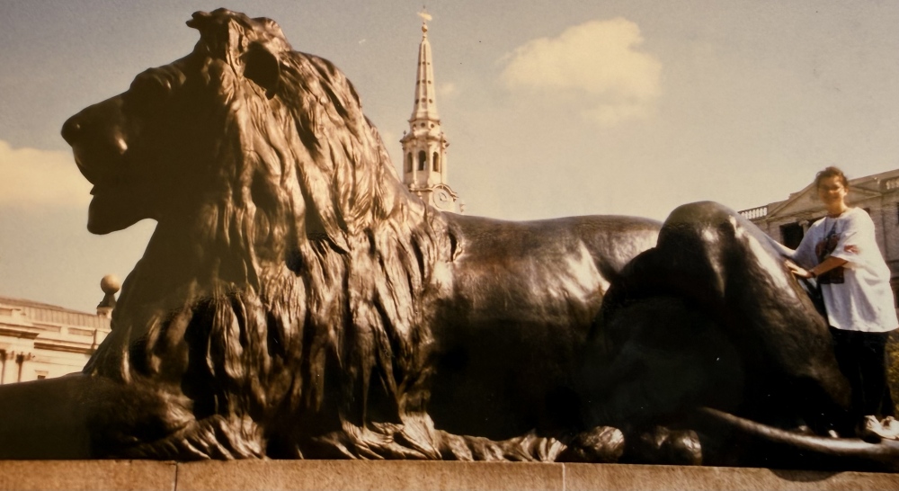 A bronze lion statue in Trafalgar Square with a church spire behind it.